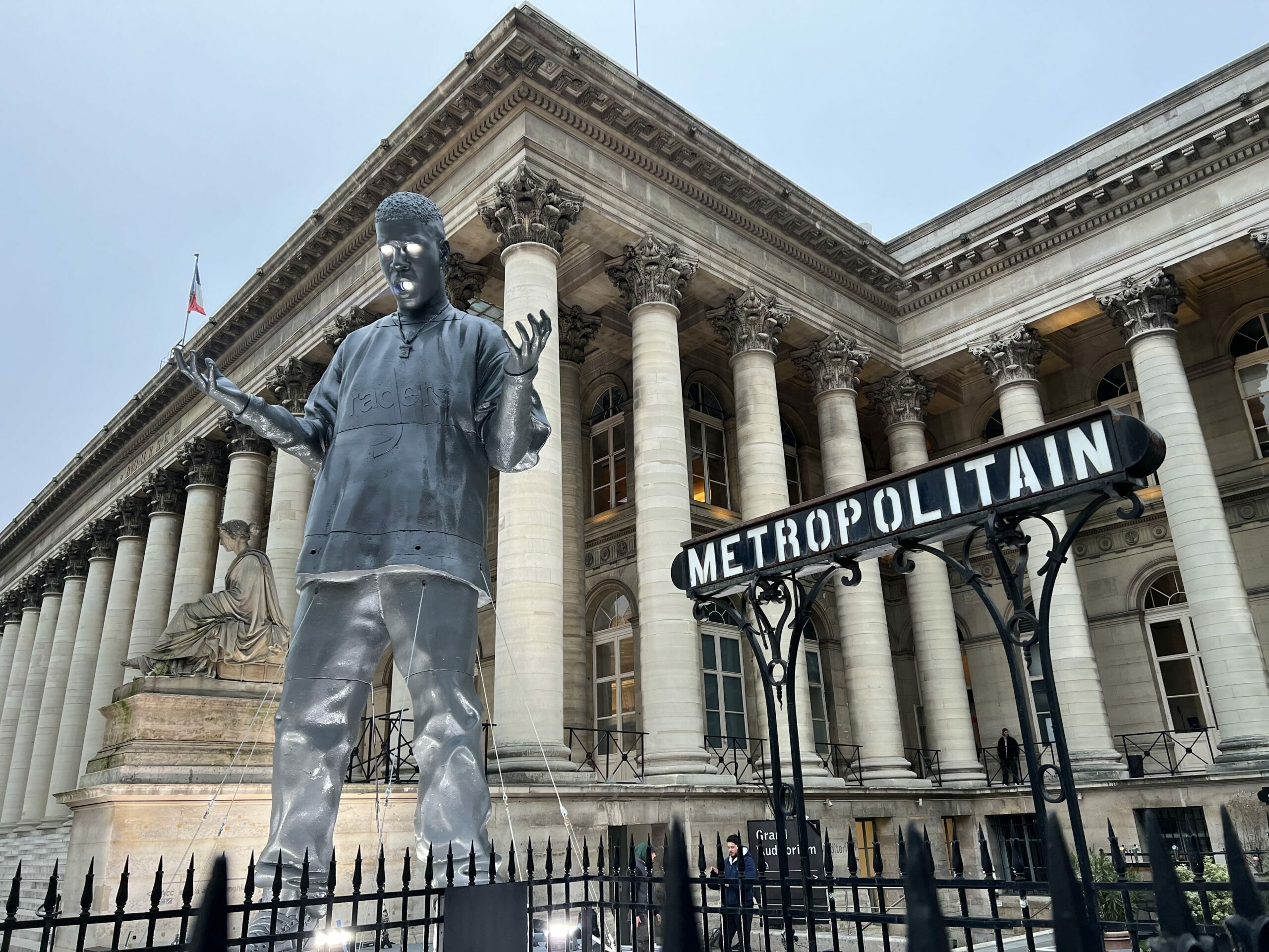 "Statue moderne devant le Palais Brongniart à Paris, avec une enseigne de métro parisien en premier plan."